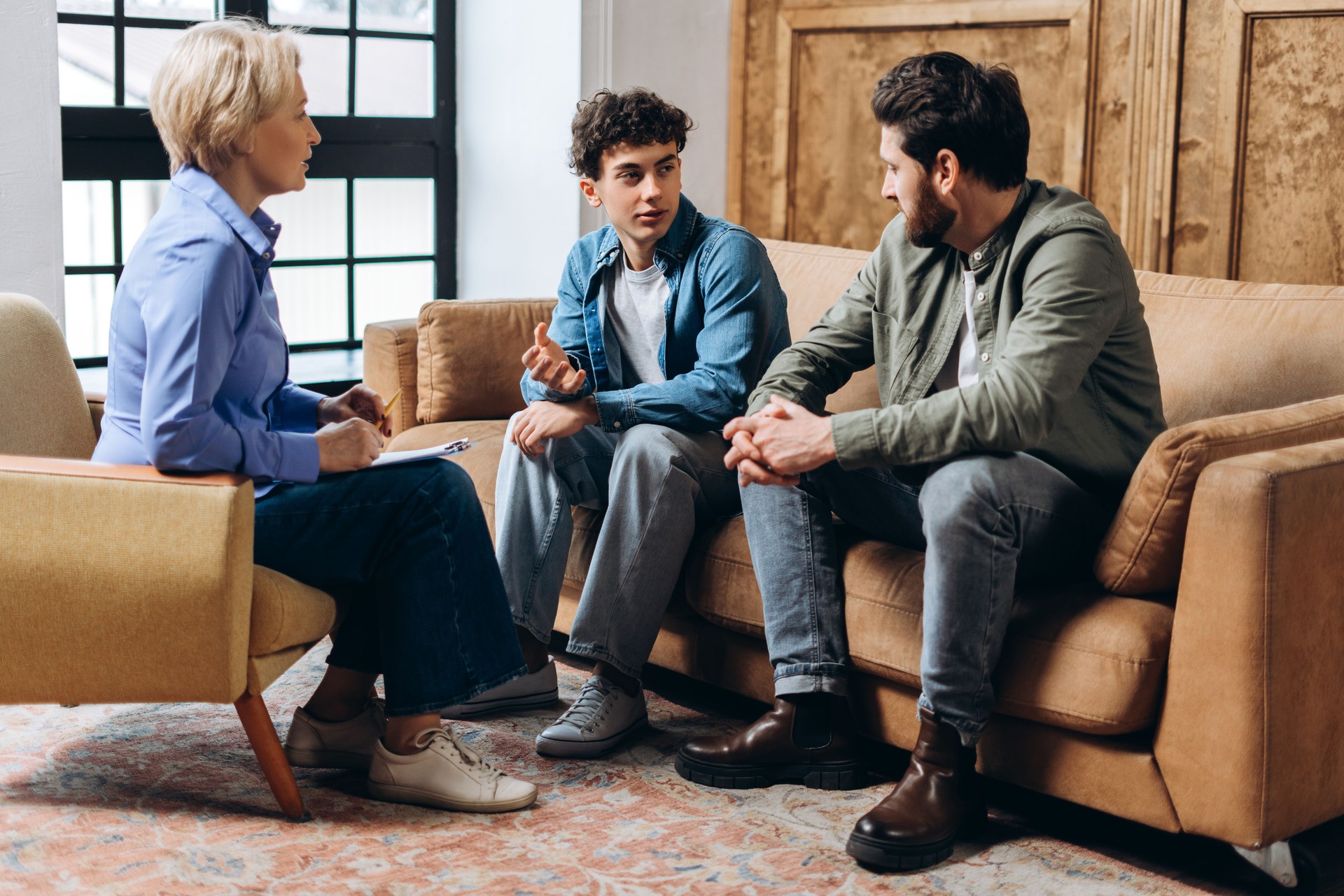 Father and teenage son talking with female psychologist sitting on sofa in the office
